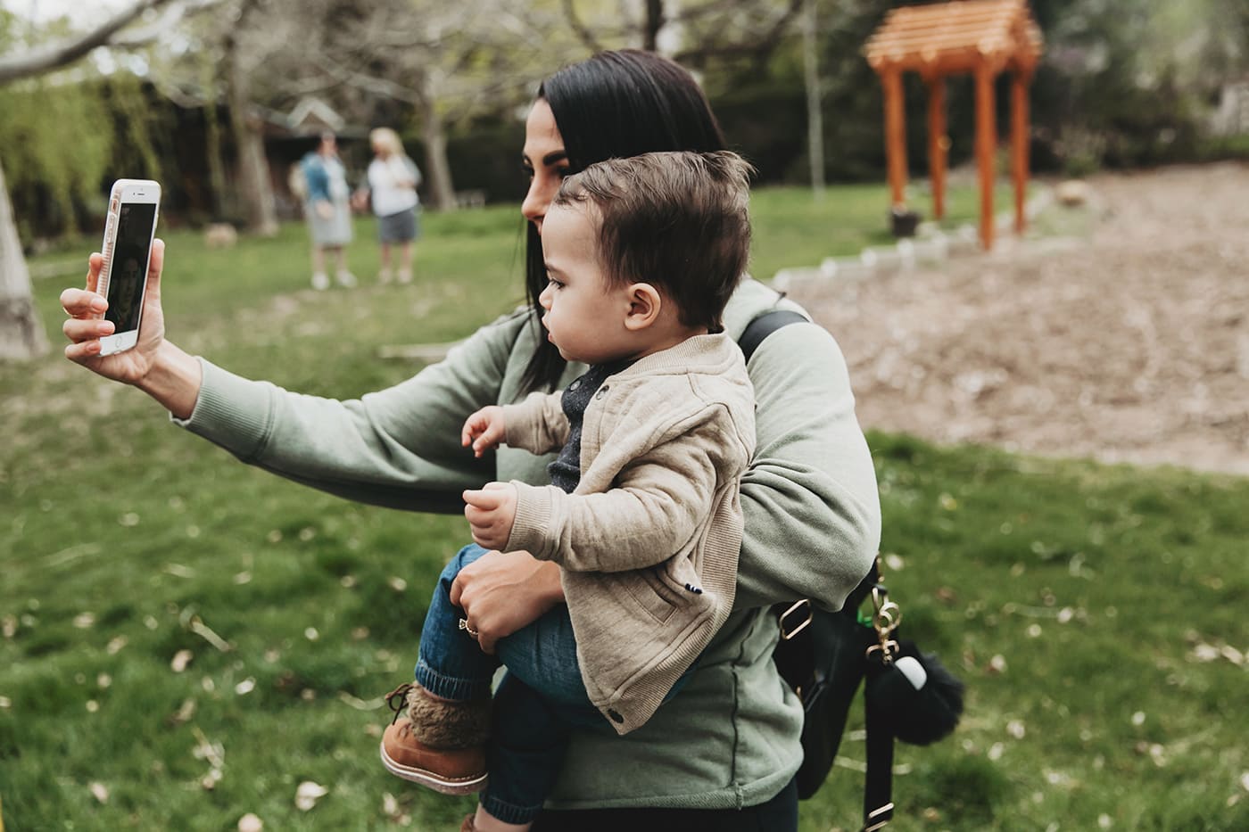 Mutter hält Kleinkind in den Armen, während sie ein Selfie zusammen in einem Park machen